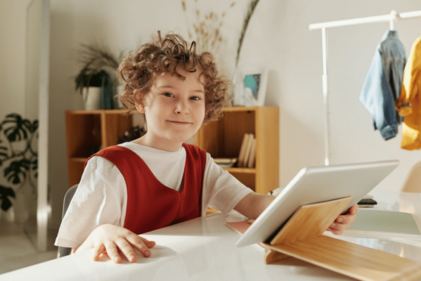 A child sitting down at a desk is holding a silver tablet and smiling while looking at the camera