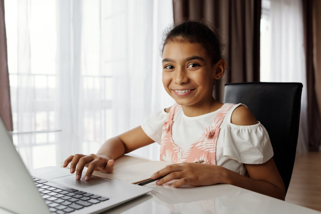 A girl wearing a white and pink top using a laptop while looking ahead at the camera and smiling